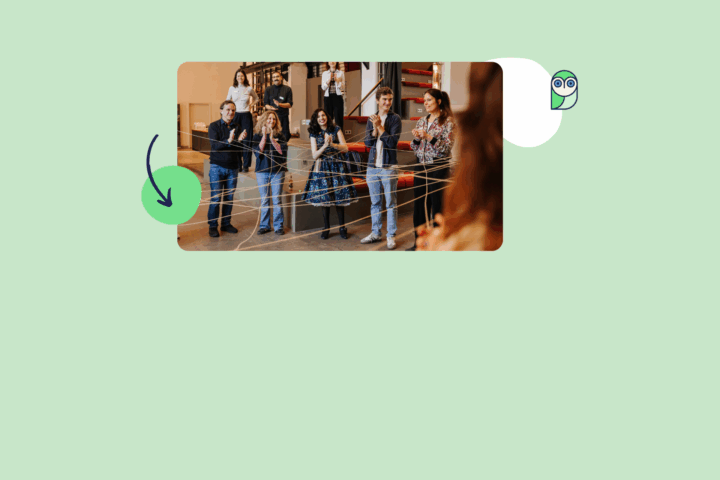 Eine Gruppe von Menschen steht in einem Raum, der mit Holzregalen und einer Treppe im Hintergrund ausgestattet ist. Die Personen sind in einem Halbkreis angeordnet und blicken auf eine Person im Vordergrund, die mit dem Rücken zur Kamera steht. Zwischen ihnen sind mehrere gelbe Fäden gespannt, die sich kreuzen und ein Netz bilden. Die Personen tragen unterschiedliche Kleidung, darunter Jeans, Röcke und Pullover.