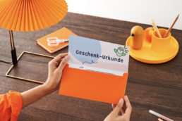 Mensch öffnet einen Briefumschlag, dort ist eine Urkunde mit dem Titel: Geschenk:Urkunde