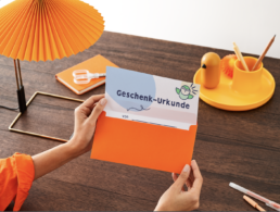 Mensch öffnet einen Briefumschlag, dort ist eine Urkunde mit dem Titel: Geschenk:Urkunde