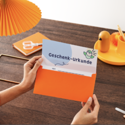 Mensch öffnet einen Briefumschlag, dort ist eine Urkunde mit dem Titel: Geschenk:Urkunde
